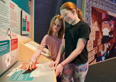 Two young girls with red hair stand in front of an interactive table at a museum exhibition and move magnets around.