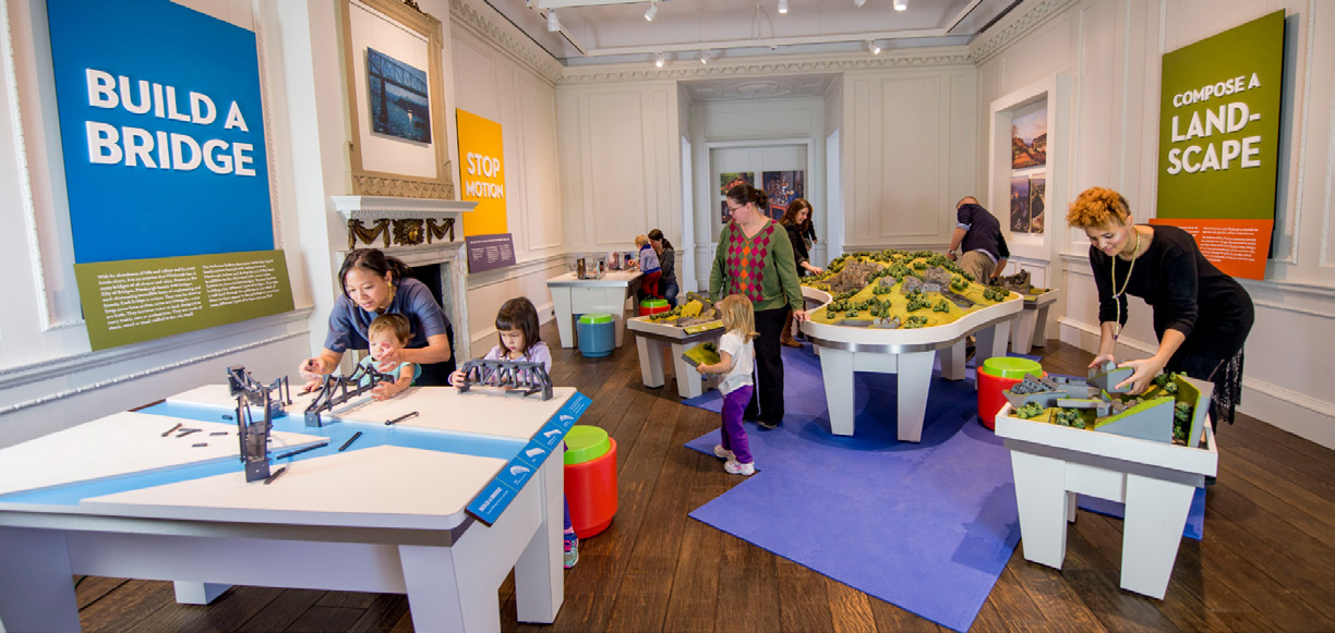 Adults and children in an interactive museum space that includes a number of tables with blocks and models for children to create and build.