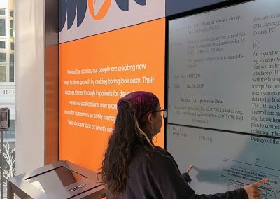 A person with eyeglasses and long hair stands in front of large interactive screen and gestures with their hands.