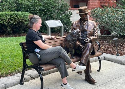A woman with a black shirt and gray pants sits on a bench outside next to a bronze sculpture of a man and a dog.