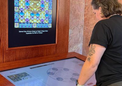 A person leans on a wooden table that encapsulates a digital interactive that guides you through designing a stained-glass window.