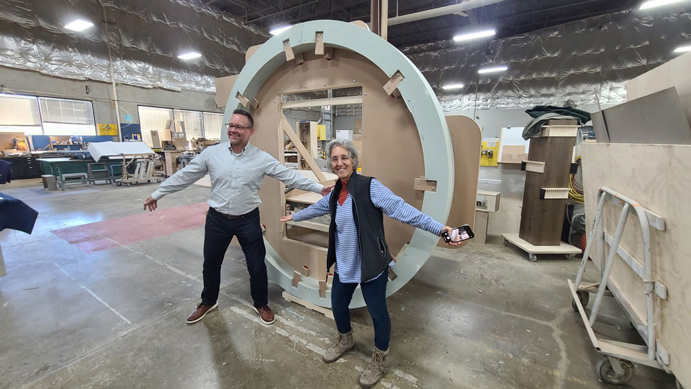 Two people spread their arms and smile as they stand in front of a large circular model in a fabrication shop.