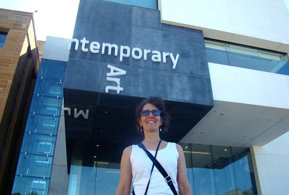 A woman wears sunglasses and a white tank top as she walks confidently outside an art museum.