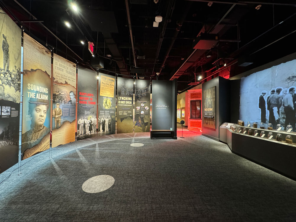 A gallery space with dark carpet and a ring of colorful banners featuring historic newspaper articles and faces.
