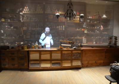 A view into an old general store in a museum. A virtual docent, dressed as a shopkeeper, appears on a screen.