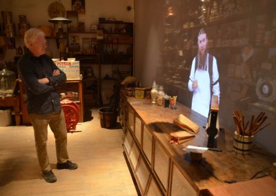 A man stands in a museum that is set up like an old general store. He watches a video of a man who is a shopkeeper.