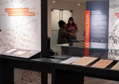 Two people are seen through the exhibit components in the gallery. In the foreground is a map, along with multiple historic documents.