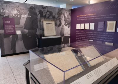 A purple wall in a museum gallery has large group of people raising their right hands in a group setting, like a naturalization ceremony.