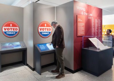 A man stands in front of a gray walls that has large I Voted stickers. He plays with a digital interactive.